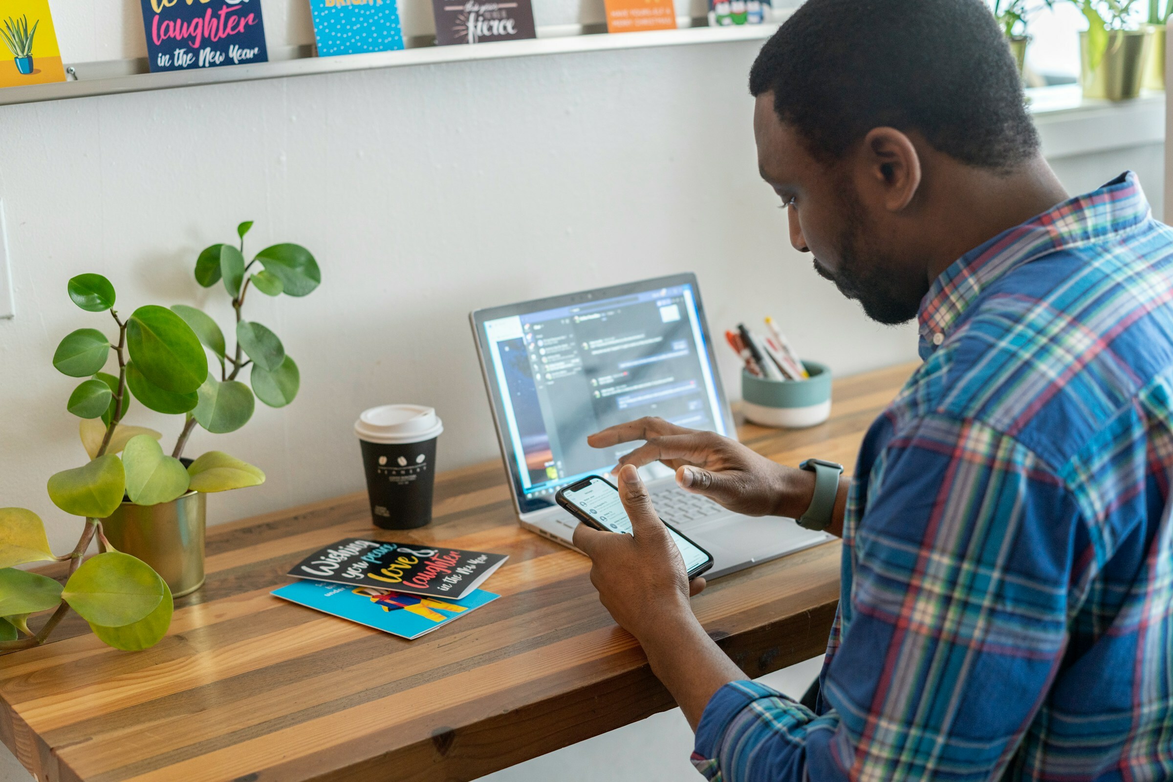 A man is sitting at a table and scrolling through a smart phone. On the table there are a tablet, a cup of coffee and a plant.