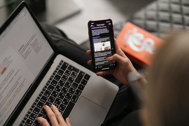 A person is scrolling through the news on a smart phone in their right hand and is navigating a laptop on their lap with their left hand.